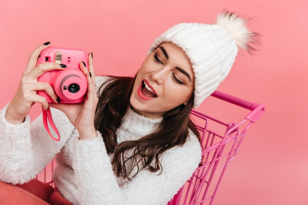 Fascinated brunette in knitted hat makes photo on camera instax. Portrait of young girl in pink tro